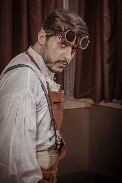 young man dressed in clothes in the style of steampunk, posing in the interior against the background of large windows