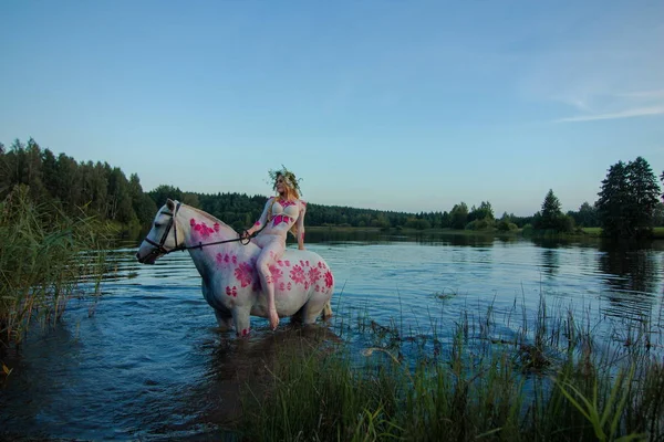 Blonde girl in white and red bodypain jumps  with a horse in the lake at sunset