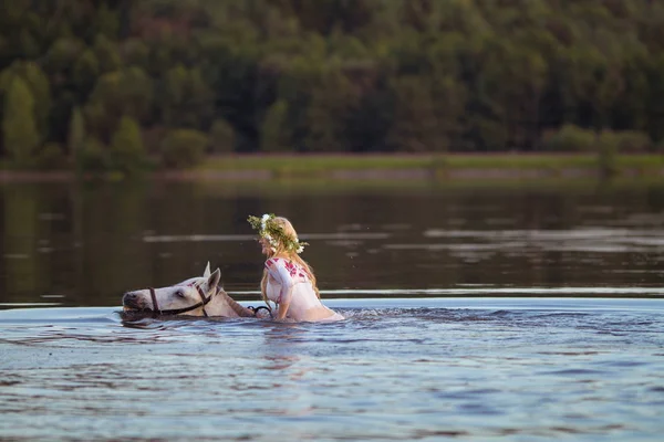 Blonde girl in white and red bodypain jumps  with a horse in the lake at sunset