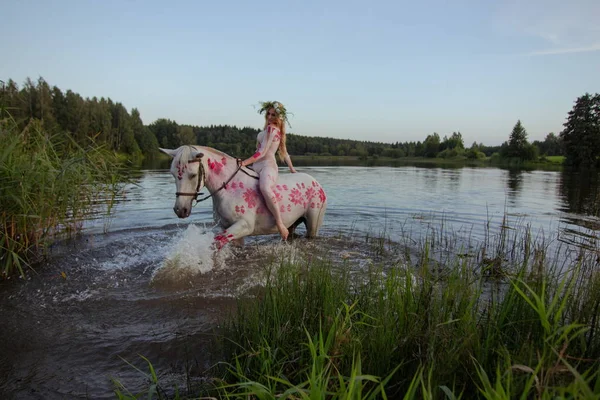 Blonde girl in white and red bodypain jumps  with a horse in the lake at sunset