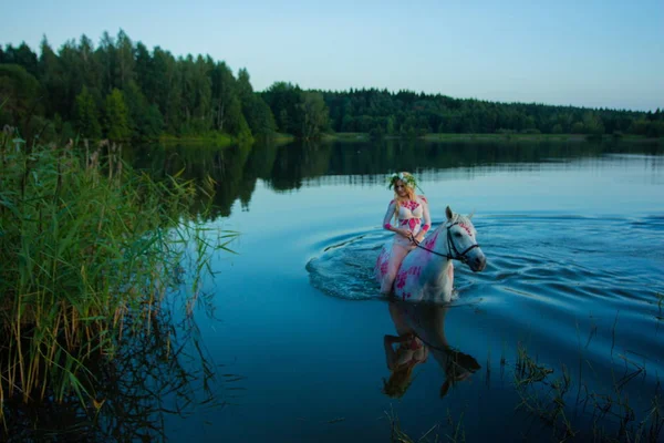 Blonde girl in white and red bodypain jumps  with a horse in the lake at sunset