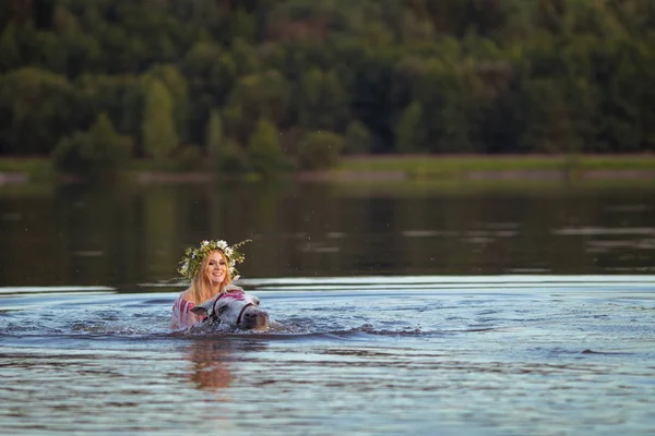 Blonde girl in white and red bodypain jumps  with a horse in the lake at sunset