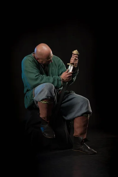 Portrait of a brutal bald man viking in linen clothes with a sword in hands posing against a black background. Early medieval period