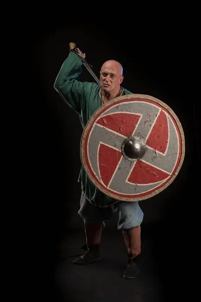 Portrait of a brutal bald man viking in linen clothes with a sword in hands posing against a black background. Early medieval period
