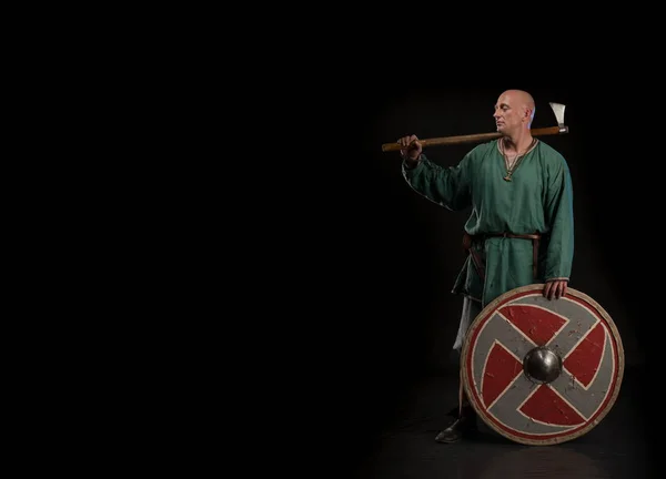 Portrait of a brutal bald man viking in linen clothes with a sword in hands posing against a black background. Early medieval period