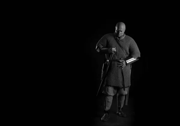 Portrait of a brutal bald man viking in linen clothes with a sword in hands posing against a black background. Early medieval period