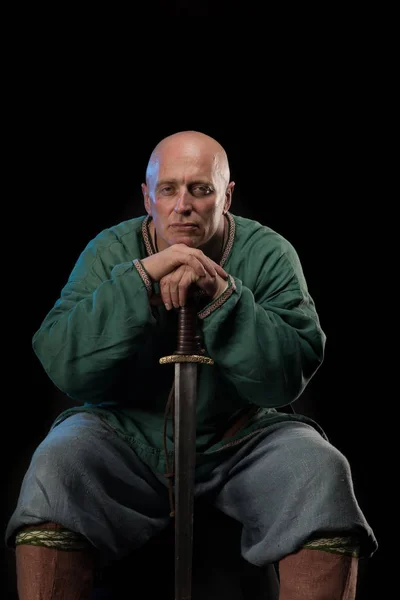 Portrait of a brutal bald man viking in linen clothes with a sword in hands posing against a black background. Early medieval period