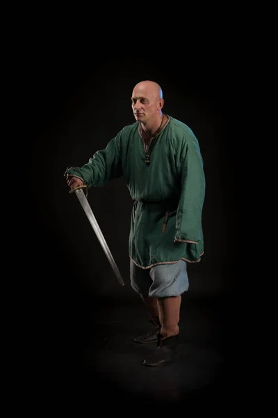 Portrait of a brutal bald man viking in linen clothes with a sword in hands posing against a black background. Early medieval period