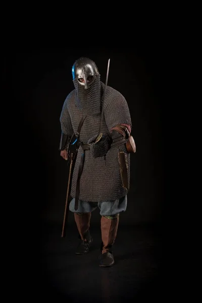 Portrait of a brutal viking in a battle mail with a helmet on his head and with a sword in his hands posing against a black background. Early medieval period