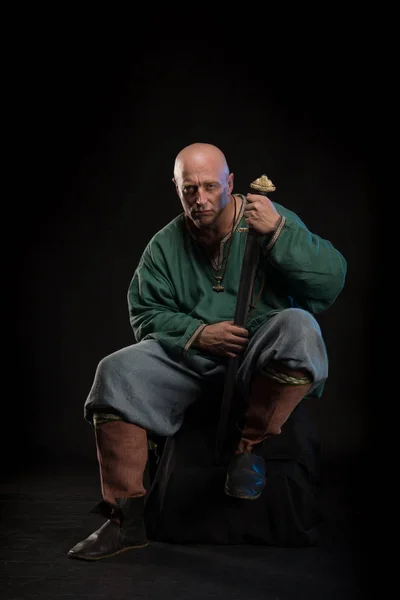 Portrait of a brutal bald man viking in linen clothes with a sword in hands posing against a black background. Early medieval period