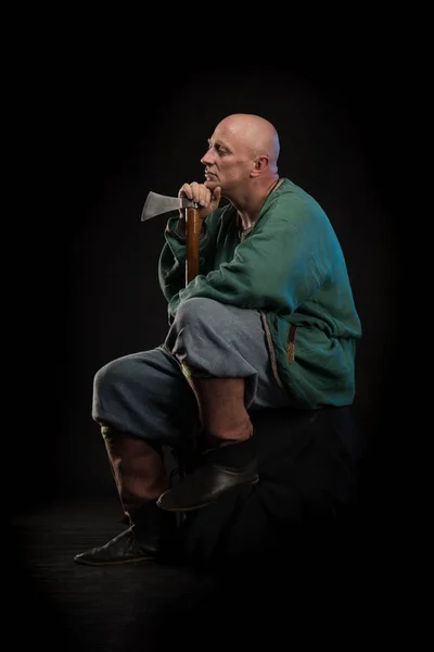 Portrait of a brutal bald man viking in linen clothes with a sword in hands posing against a black background. Early medieval period