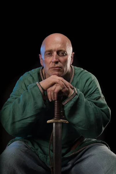 Portrait of a brutal bald man viking in linen clothes with a sword in hands posing against a black background. Early medieval period