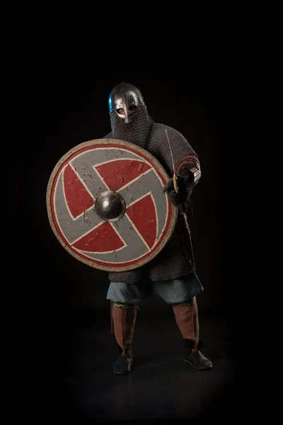 Portrait of a brutal man viking in linen clothes with shield and sword in hands posing against a black background. Early medieval period