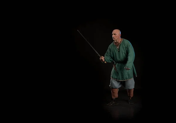 Portrait of a brutal bald man viking in linen clothes with a sword in hands posing against a black background. Early medieval period