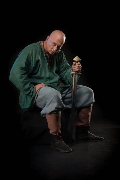 Portrait of a brutal bald man viking in linen clothes with a sword in hands posing against a black background. Early medieval period