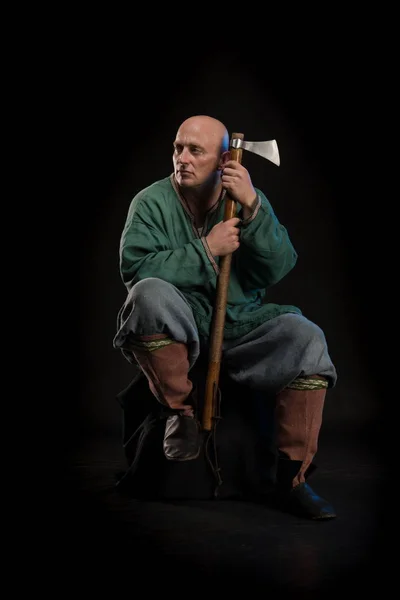 Portrait of a brutal bald man viking in linen clothes with a sword in hands posing against a black background. Early medieval period