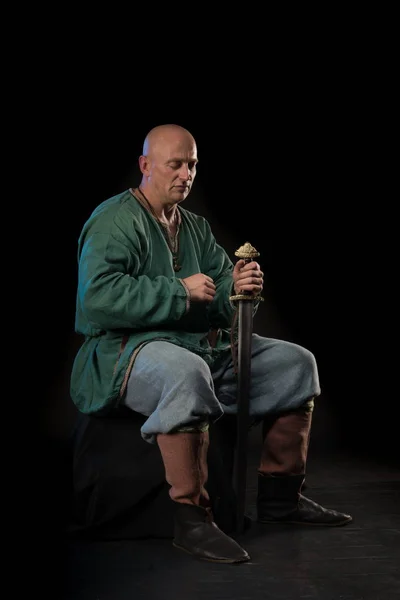 Portrait of a brutal bald man viking in linen clothes with a sword in hands posing against a black background. Early medieval period