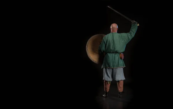 Portrait of a brutal bald man viking in linen clothes with a sword in hands posing against a black background. Early medieval period