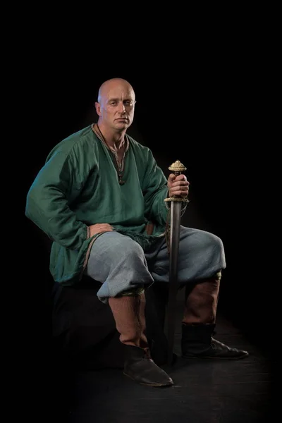 Portrait of a brutal bald man viking in linen clothes with a sword in hands posing against a black background. Early medieval period