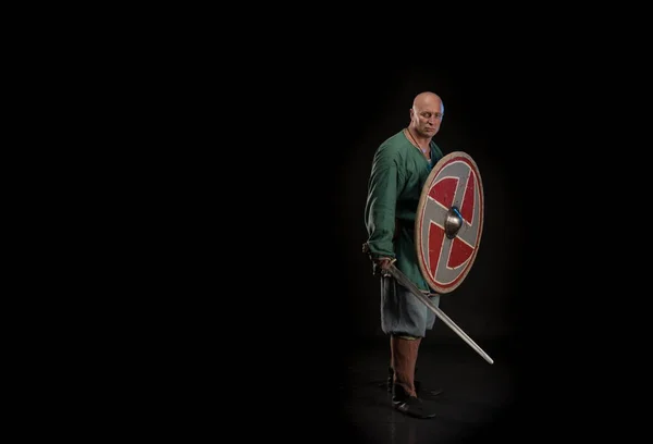 Portrait of a brutal bald man viking in linen clothes with a sword in hands posing against a black background. Early medieval period