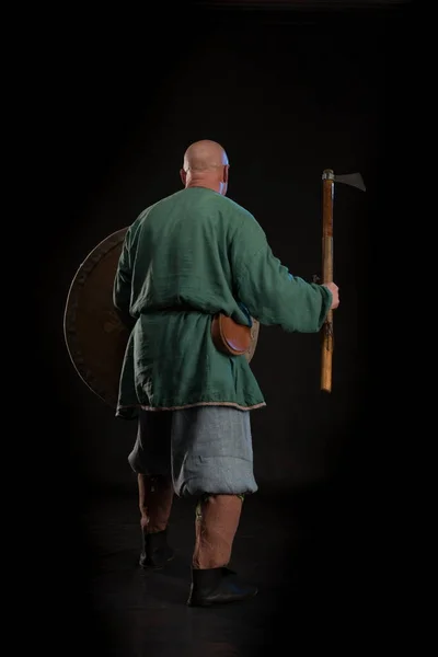 Portrait of a brutal bald man viking in linen clothes with a sword in hands posing against a black background. Early medieval period