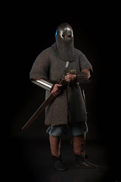 Portrait of a brutal Viking in a military mail with a helmet on his head holding sword on a black background. Early medieval period