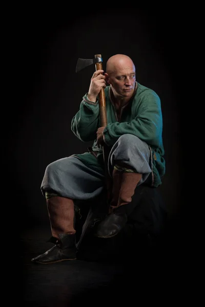 Portrait of a brutal bald man viking in linen clothes with a sword in hands posing against a black background. Early medieval period