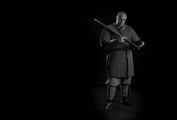 Portrait of a brutal bald man viking in linen clothes with a sword in hands posing against a black background. Early medieval period