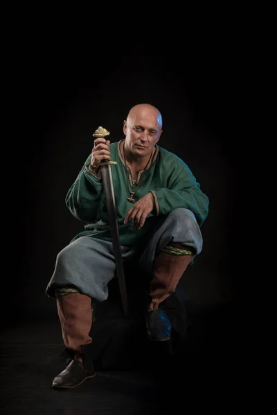 Portrait of a brutal bald man viking in linen clothes with a sword in hands posing against a black background. Early medieval period