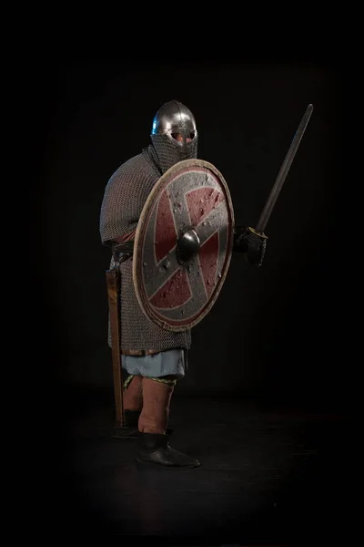 Portrait of a brutal man viking in linen clothes with shield and sword in hands posing against a black background. Early medieval period