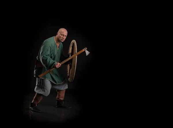 Portrait of a brutal bald man viking in linen clothes with a sword in hands posing against a black background. Early medieval period