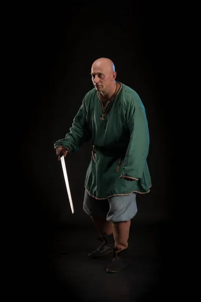 Portrait of a brutal bald man viking in linen clothes with a sword in hands posing against a black background. Early medieval period