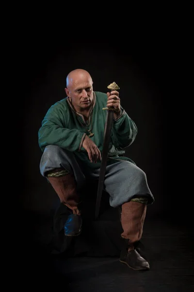 Portrait of a brutal bald man viking in linen clothes with a sword in hands posing against a black background. Early medieval period