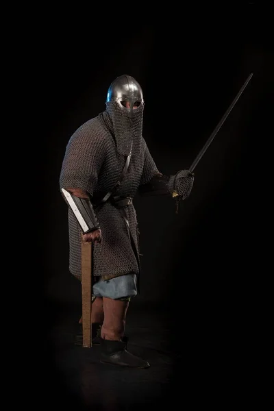 Portrait of a brutal viking in a battle mail with a helmet on his head and with a sword in his hands posing against a black background. Early medieval period