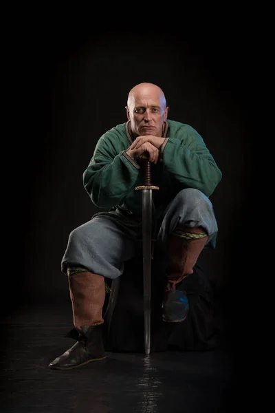 Portrait of a brutal bald man viking in linen clothes with a sword in hands posing against a black background. Early medieval period