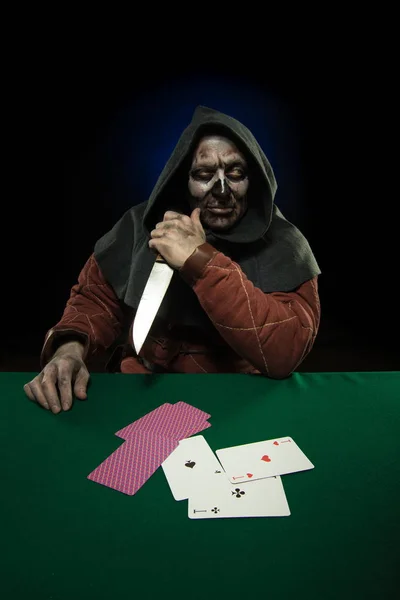 male actor in the costume of a medieval inquisitor playing poker at a card table with a green cloth