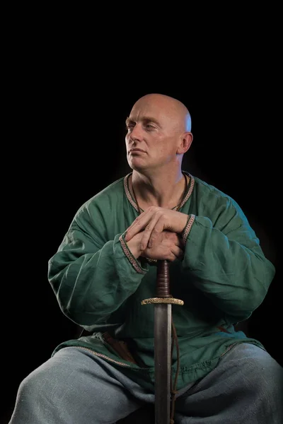 Portrait of a brutal bald man viking in linen clothes with a sword in hands posing against a black background. Early medieval period