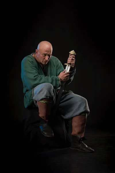 Portrait of a brutal bald man viking in linen clothes with a sword in hands posing against a black background. Early medieval period