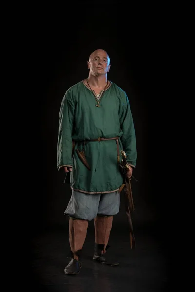 Portrait of a brutal bald man viking in linen clothes with a sword in hands posing against a black background. Early medieval period