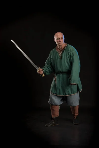 Portrait of a brutal bald man viking in linen clothes with a sword in hands posing against a black background. Early medieval period