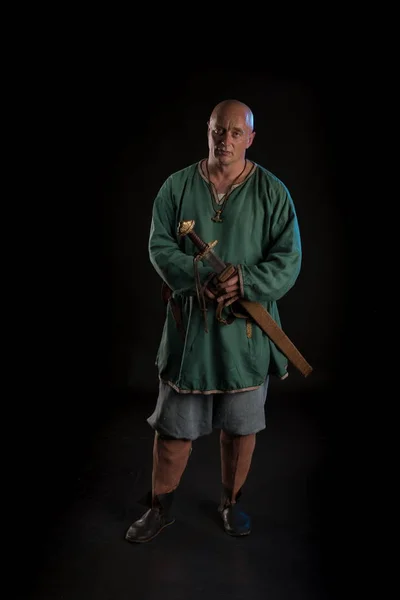 Portrait of a brutal bald man viking in linen clothes with a sword in hands posing against a black background. Early medieval period