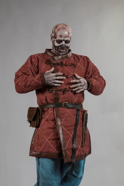 Male actor in zombie viking make up and costume posing with weapon on grey background