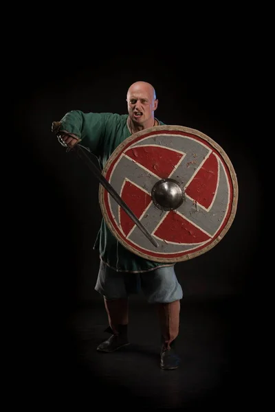 Portrait of a brutal bald man viking in linen clothes with a sword in hands posing against a black background. Early medieval period