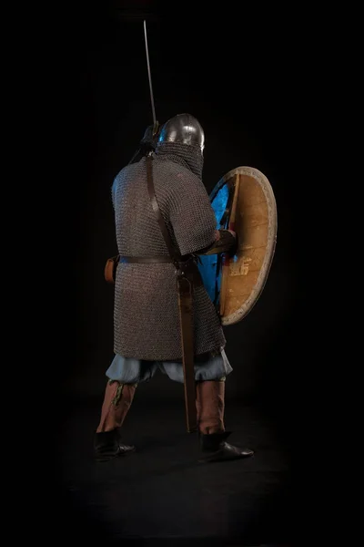 Portrait of a brutal man viking in linen clothes with shield and sword in hands posing against a black background. Early medieval period