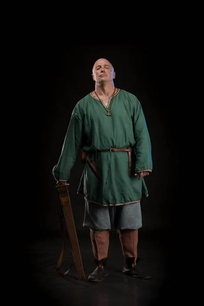 Portrait of a brutal bald man viking in linen clothes with a sword in hands posing against a black background. Early medieval period