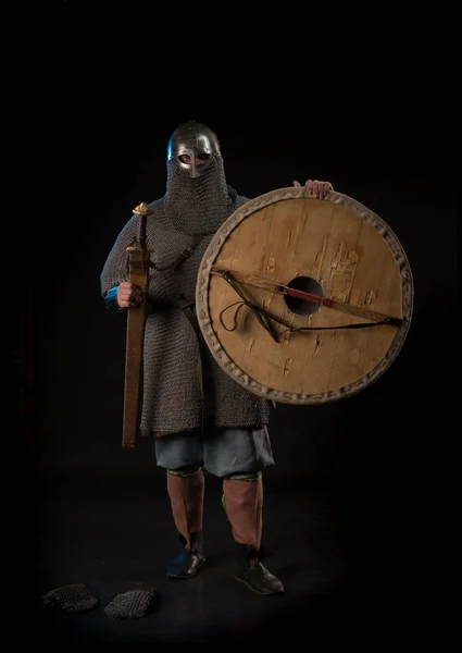 Portrait of a brutal viking in a chain armor with a helmet, shield and sword posing against a black background. Early medieval period.