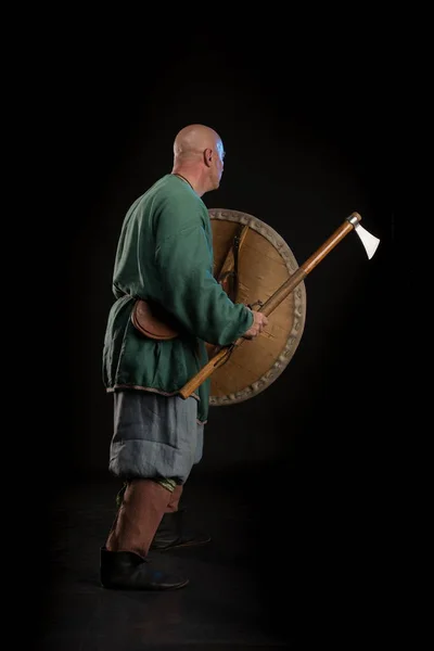 Portrait of a brutal bald man viking in linen clothes with a sword in hands posing against a black background. Early medieval period