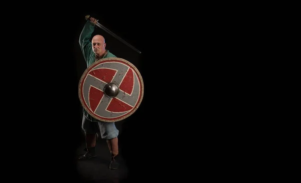 Portrait of a brutal bald man viking in linen clothes with a sword in hands posing against a black background. Early medieval period