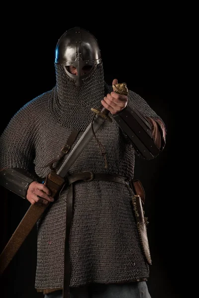 Portrait of a brutal Viking in a military mail with a helmet on his head holding sword on a black background. Early medieval period