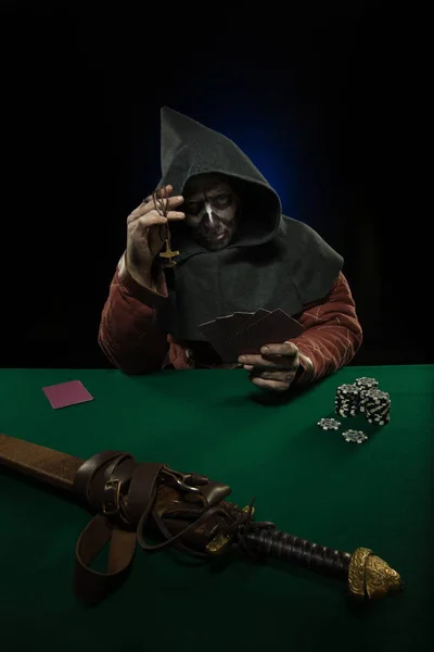 male actor in the costume of a medieval inquisitor playing poker at a card table with a green cloth